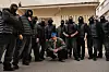 A man wearing a blue hat and grey leather jacket is squatting among a group of men in all black, with their faces covered.