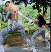Two dancers in leopard-print bodysuits perform energetic poses outdoors near a low wall and trees.