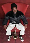 An image of Linda Sikhakhane sitting on a red chair and placing his saxophone between his legs.