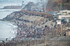 Crowds fill a coastal promenade beside the ocean, stretching along a breakwater under a hazy sky.