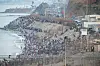 Crowds fill a coastal promenade beside the ocean, stretching along a breakwater under a hazy sky.