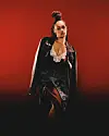 Nanette is in a black leather jacket and dark outfit, standing against a red background.