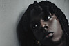 Close-up of Rema with twisted locs looking up beside a gray wall.