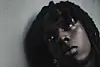 Close-up of Rema with twisted locs looking up beside a gray wall.