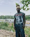 Llona is standing near a road with trees, grass, and low buildings behind him.