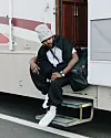 Llona, in a beanie and dark clothing, sits on the steps of a white trailer.