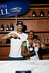 Mixologists Thandi Walton and Marcio Ramos pose at a Martell-branded bar with bottles and glasses on wooden shelves.