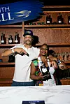 Mixologists Thandi Walton and Marcio Ramos pose at a Martell-branded bar with bottles and glasses on wooden shelves.