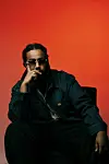 Man in sunglasses and dark jacket seated against a bright red background.