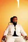 A seated man in light clothing poses against a bright orange studio backdrop.