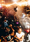 Crowded nighttime outdoor scene with people dancing under bright lights and smoke.