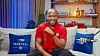 Smiling man in a red shirt sits on a sofa with Martell pillows in a studio setting.
