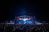 A large outdoor concert crowd watches a brightly lit stage at night on the beach in New Alamein, Egypt.