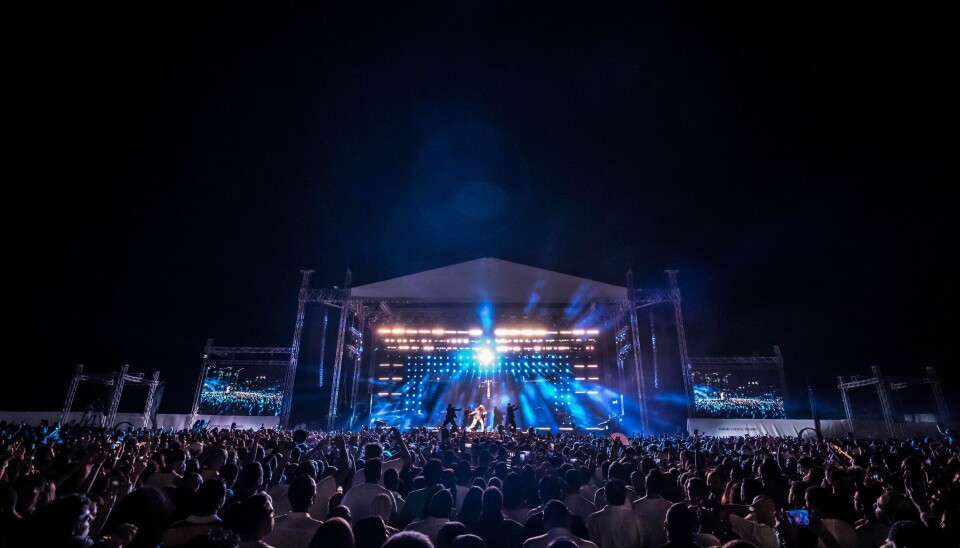 A large outdoor concert crowd watches a brightly lit stage at night on the beach in New Alamein, Egypt.