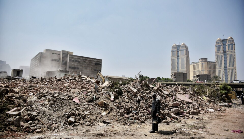 Security officer stands beside a large rubble pile in Cairo with high-rise towers in the distance.