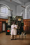 Kenya’s Fena Gitu dressed in a white coat and tan wide-leg pants before stacked vintage speakers in a wood-paneled room