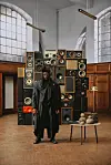 Cameroon’s Tenor dressed in black and standing before stacked vintage speakers in a wood-paneled room