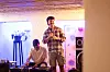 A man wearing a chequered shirt is standing in front of a crowd, laughing and speaking into a microphone while another man behind him is sitting by a mixing deck, also smiling.