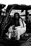 Black-and-white portrait of Adey seated inside a utility vehicle, wearing a white embroidered dress and black sneakers.