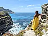 Jalisa Whitley, in a flowing yellow dress, stands on a rocky cliff overlooking the blue ocean and coastline.