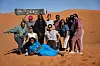 A group of women poses together on orange sand dunes in the Moroccan desert, with one traveler holding a sandboard overhead and another reclining in front of the group.