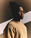 Manana in glasses and a beige sweater, set against a dark background with diagonal lighting.