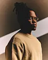 Manana in glasses and a beige sweater, set against a dark background with diagonal lighting.