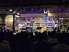 Audience silhouettes watch musicians performing on a small stage in a book-lined venue.