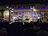 Audience silhouettes watch musicians performing on a small stage in a book-lined venue.