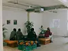 People seated indoors watch a projected presentation in a room with plants and produce crates.