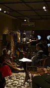 People sit and stand in a dim room beside a clothing rack and patterned floor.