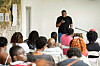 Speaker with a microphone addresses a seated audience in a bright room.