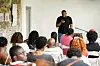 Speaker with a microphone addresses a seated audience in a bright room.