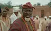 Ogogo in traditional clothing and coral beads stands before a crowd outdoors.