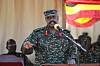 Ugandan military officer in camouflage speaks into a microphone before a flag backdrop.