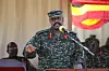 Ugandan military officer in camouflage speaks into a microphone before a flag backdrop.