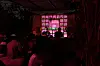 A DJ is playing for a full room in a window that is lit up with pink light. In the room behind him, the wall is decorated with different paraphernalia and a poster that reads Yellow Tape.
