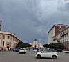 Cars cross a city intersection beneath dark storm clouds and rows of historic buildings.