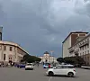 Cars cross a city intersection beneath dark storm clouds and rows of historic buildings.