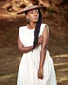 Woman in a white dress and woven hat standing among trees with a long braid over one shoulder.