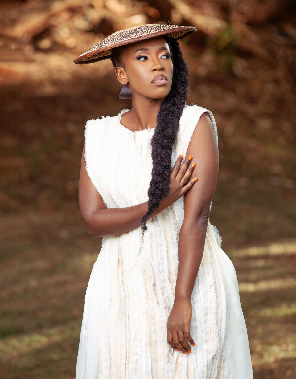 Woman in a white dress and woven hat standing among trees with a long braid over one shoulder.