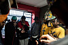 Group of rappers gathered indoors near a barred window, with cables and equipment in the foreground, mics in hand.