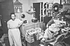 Two women work in a densely furnished vintage workshop filled with tools, equipment, and shelves.