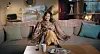 Yousra is sitting on a beige sofa, wearing a golden skirt and an animal cheeta print blouse, smiling mischievously and looking pleased with herself.