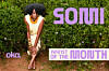 Woman in a cream dress poses before a hedge with purple promotional lettering.