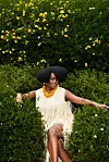 Somi is dressed in a cream-fringed dress, posing amid dense greenery and colorful flowers.