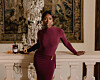 Ivie Ani stands in a burgundy dress beside an ornate white mantel in an elegant room.