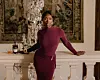 Ivie Ani stands in a burgundy dress beside an ornate white mantel in an elegant room.
