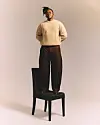 Manana is wearing a beige sweater, standing on a black chair in a softly lit studio.