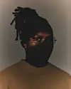 Close-up portrait of a Manana with locs and glasses wearing a tan sweater in low, warm light.